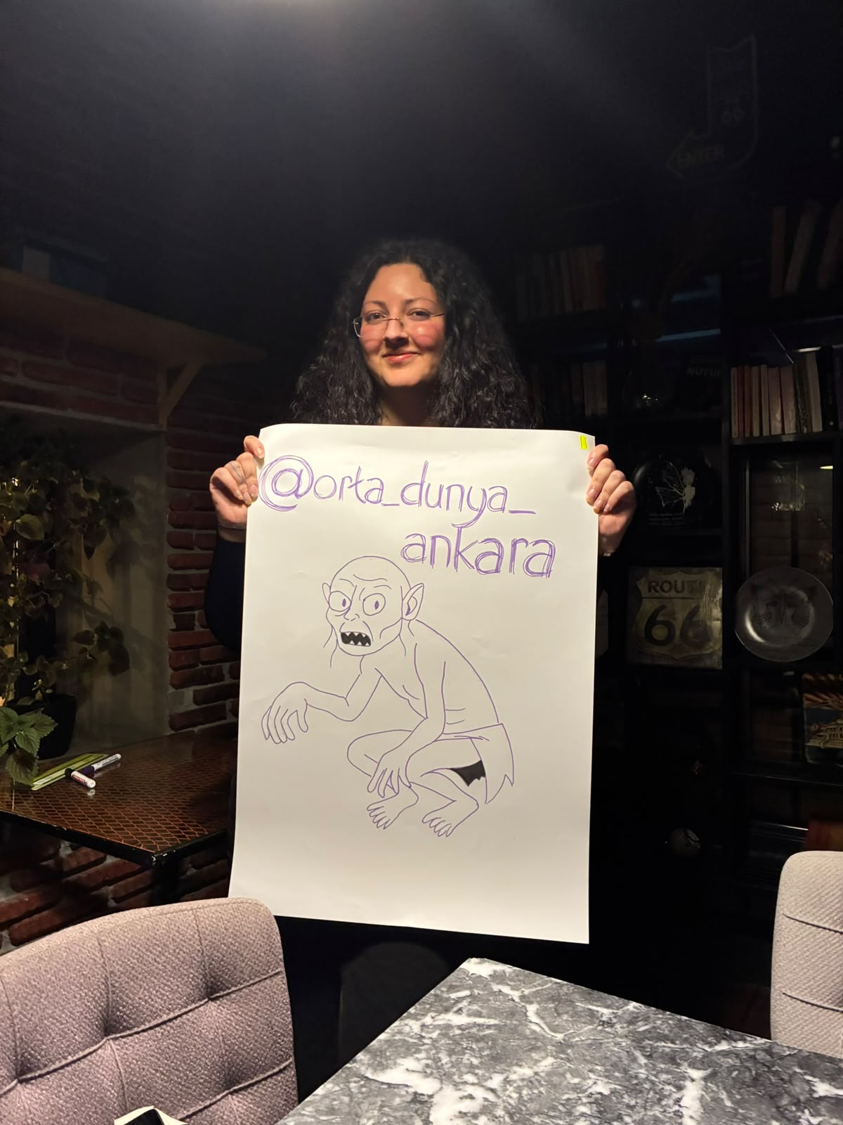 Bir katılımcı, Gollum çizimi ve @orta_dunya_ankara yazısı olan afişi tutuyor.
