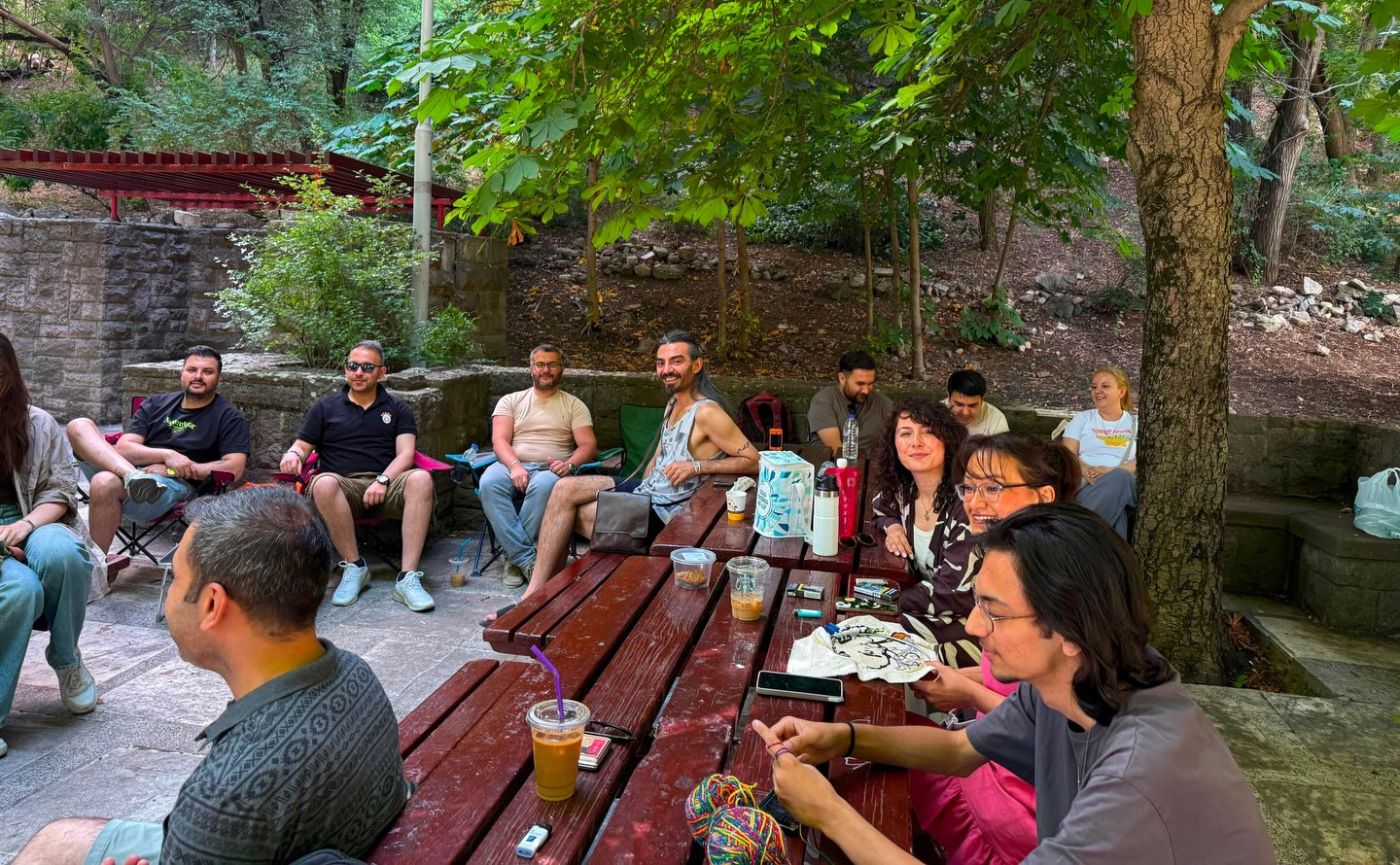 Ağaçların gölgesinde masa ve sandalyelerde sohbet eden topluluk.